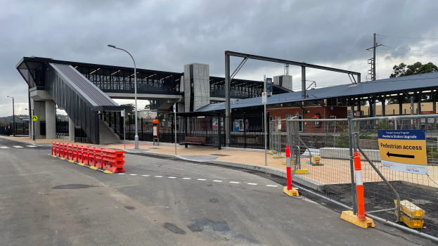 Ein Bahnhofsbild mit einer Brücke im Hintergrund, Straßenlaternen, Strommasten, Stromkabel, Bäume, Gebäude, Fahrzeuge auf der Straße, Absperrpoller, Schilder, Bänke und einen bewölkten Himmel.
