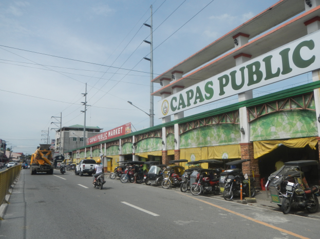 Eine belebte Stadtstraße mit Fahrzeugen, einem Geländer auf der linken Seite, Gebäuden, Pfählen, Lampen, Schildern und einigen Menschen, unter einem bewölkten Himmel, mit einem Gebäude mit der Aufschrift "Capas Public".
