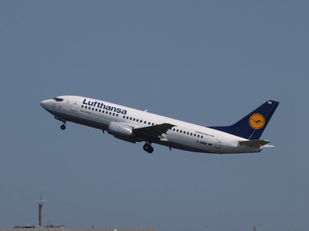 Lufthansa Airbus A320-200-Flugzeug im Flug vor einem klaren blauen Himmel, mit Gebäuden und Polen unten sichtbar.