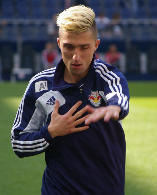 Ein junger Mann in einem blauen T-Shirt steht auf einem Fußballfeld mit ausgestreckten Armen und sieht entschlossen aus, während er als Flügelspieler mit einem unscharfen Hintergrund dargestellt wird.