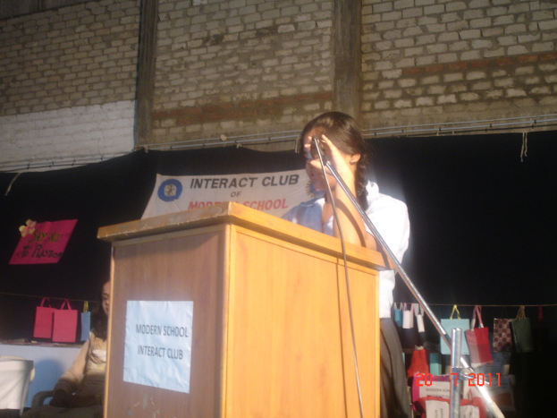 Ein Mädchen in einem weißen Hemd steht neben einem Rednerpult in einem Konferenzraum, mit einem Mikrofonständer rechts daneben, einem weißen Banner an einer Wand hinter ihm und Taschen, die an einem Faden rechts hängen.