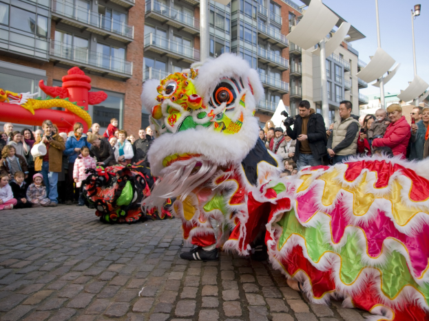 Lebendige Feier des chinesischen Neujahrs in Amsterdam mit einem Löwen tanzen im Vordergrund und einer Menschenmenge drumherum, einige halten Kameras, vor einem Hintergrund von Gebäuden, Laternenmasten und einem klaren blauen Himmel.