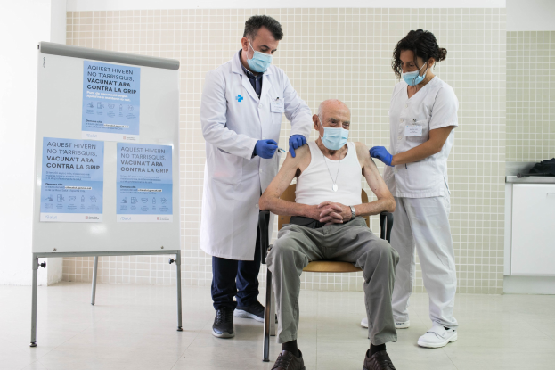 Ein älterer Mann sitzt in einem Stuhl und erhält eine Impfung von zwei maskierten und handschuhgetragenen Ärzten in einer Krankenhausumgebung, mit einer Texttafel links und medizinischen Gegenständen auf einer Plattform rechts.