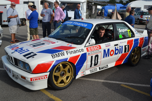 Ein weißes, blaues, rotes und schwarzes BMW M3 Motorsport Auto steht auf einem Parkplatz mit einer Person auf dem Fahrersitz, umgeben von einer Gruppe von Menschen, Zelten, Bäumen und einem klaren blauen Himmel.