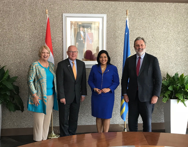 Eine Gruppe strahlender Menschen um einen Tisch mit Papieren herumstehend, umgeben von Fahnen, Topfpflanzen und einem Foto auf dem Wand, die Unterzeichnung eines Memorandums zwischen den Vereinigten Staaten und Kanada feiernd.