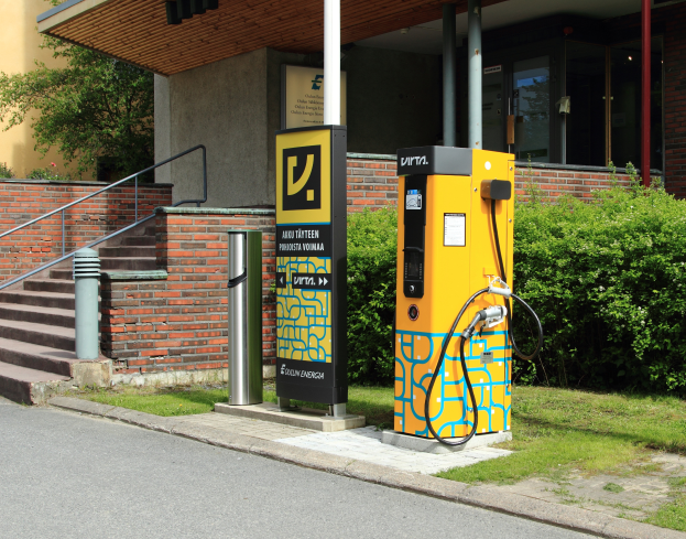Eine öffentliche Ladestation für Elektrofahrzeuge an einer Straße, umgeben von Grün und einem Gebäude mit Fenstern und Säulen, mit Treppen und Geländern, die hinaufführen.