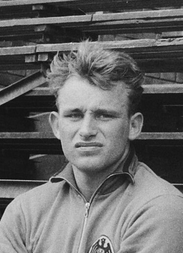 Schwarzes und weißes Foto eines Mannes in einer Jacke, der vor gestapelten Holzplanken steht, der legendäre deutsche Füßballer.