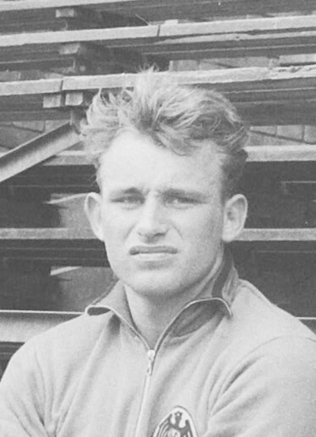 Schwarzes und weißes Foto eines Mannes in einer Jacke, der vor gestapelten Holzplanken steht, der legendäre deutsche Füßballer.