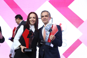 Zwei Personen auf einer Bühne, die lächeln und Pokale halten, mit einem sichtbaren Bildschirm im Hintergrund.