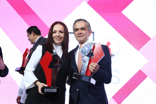 Zwei Personen auf einer Bühne, die lächeln und Pokale halten, mit einem sichtbaren Bildschirm im Hintergrund.