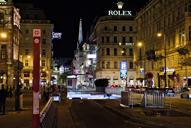 Eine nächtliche Stadtstraße mit fahrenden Autos, Fußgängern auf dem Gehweg, parkenden Fahrrädern, Straßenlaternen, hohen Gebäuden, Absperrungen, Polen und einem Rolex-Logo, das auf einem Gebäude im Hintergrund zu sehen ist.