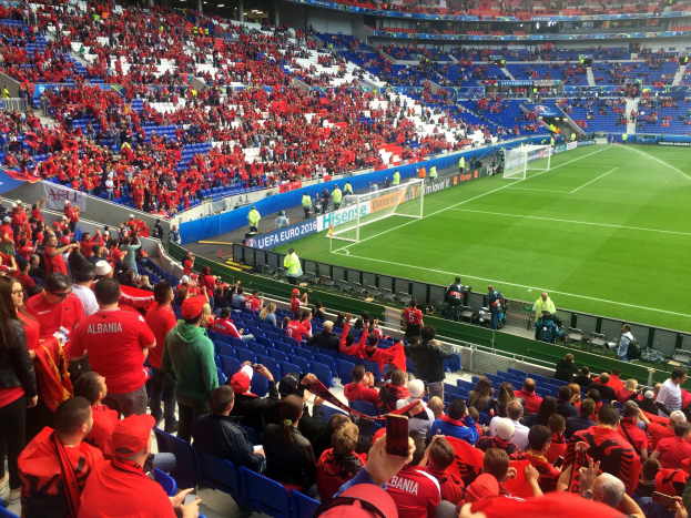 Eine große Menschenmenge in einem Stadion schaut einem Fußballspiel zu, mit Menschen, die auf Stühlen sitzen und stehen, Bannern, einem Torpfosten und Deckenlampen.