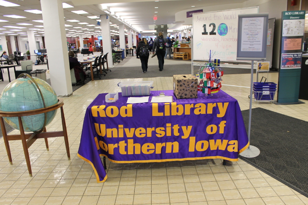 Eine Ausstellung mit einem Tisch, auf dem verschiedene Landesflaggen, eine Box, Papiere und eine Fahne stehen, eine Kugel zur Linken, ein Anschlagbrett zur Rechten mit einigen Menschen in der Nähe und mehreren Tischen und Stühlen links.