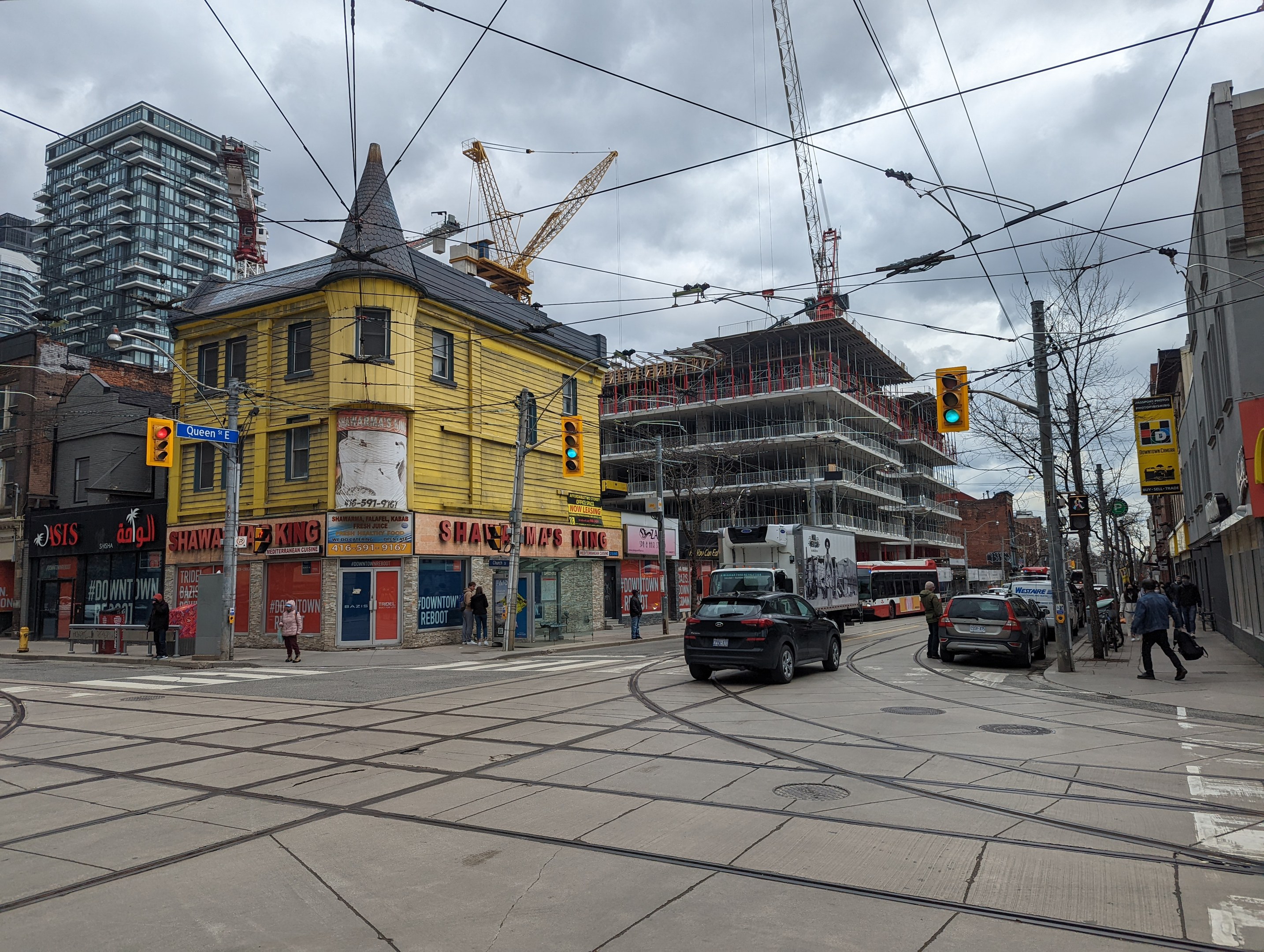 Eine Straßenszene mit einem gelben Gebäude im Bau, Autos, Fußgängern, Strommasten, Ampeln, Gebäuden, Schildern, Bäumen, Kränen und einem bewölkten Himmel.