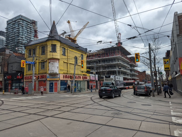 Eine Straßenszene mit einem gelben Gebäude im Bau, Autos, Fußgängern, Strommasten, Ampeln, Gebäuden, Schildern, Bäumen, Kränen und einem bewölkten Himmel.