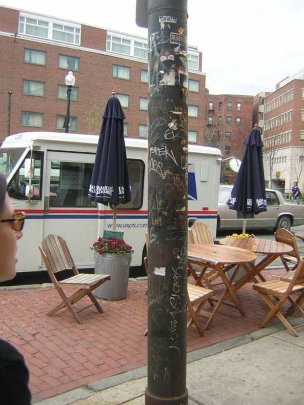 Ein Tisch und Stühle sind auf einem Gehweg aufgestellt, Autos und Lieferwagen parken auf der benachbarten Straße, und Gebäude sind im Hintergrund sichtbar.