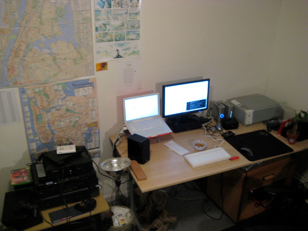 Ein Raum mit einem Tisch, auf dem ein Laptop, ein Lautsprecher, ein Monitor, eine Tastatur, ein Ball, eine Maus, ein Scanner und Papiere stehen, sowie ein weiterer Tisch mit Elektronikgeräten und Charts und Postern an der Wand dahinter.