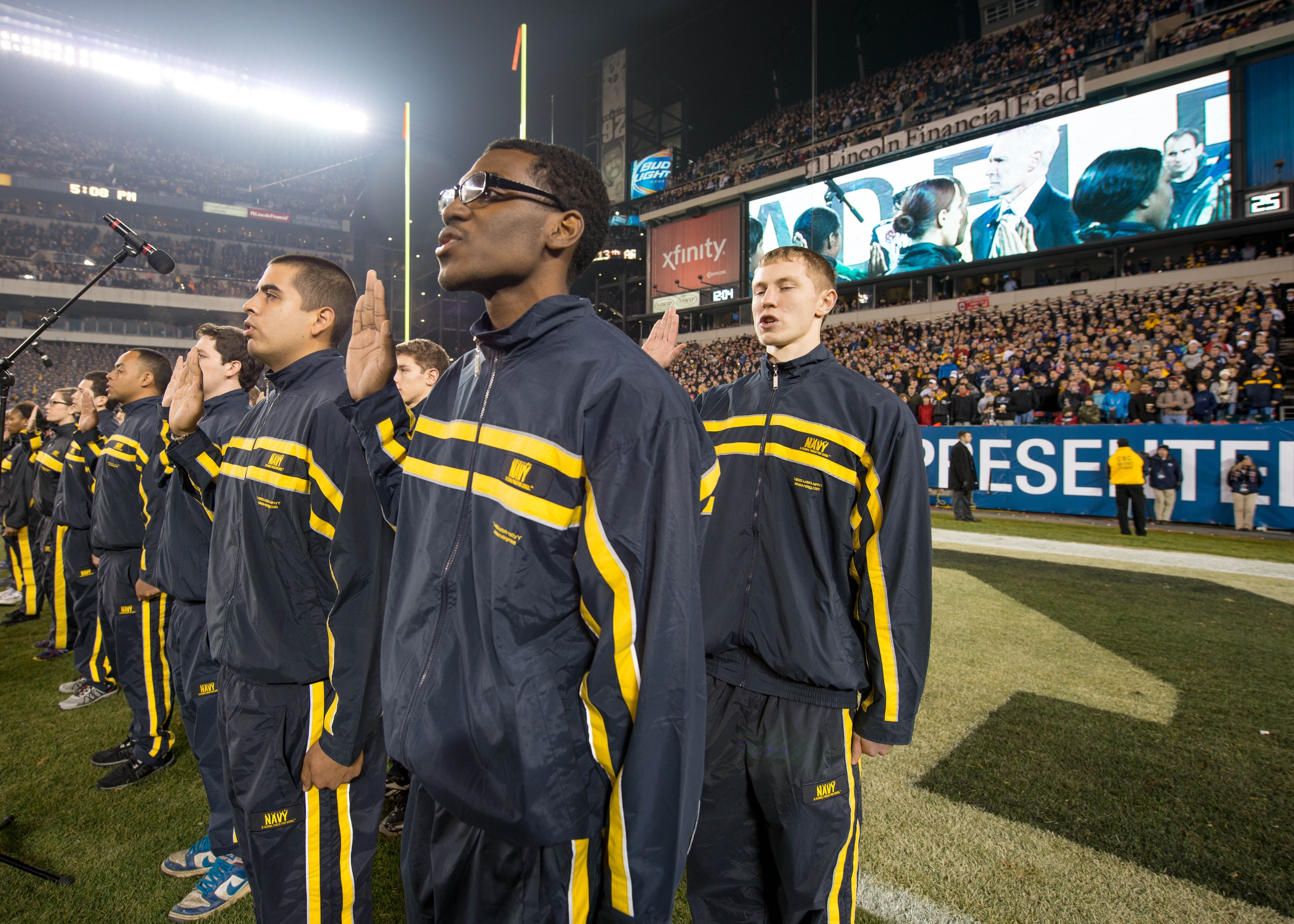 Eine Gruppe von M%C3%A4nnern in blauen und gelben Jacken steht auf einem Fu%C3%9Fballfeld, mit einem Stadionpublikum, Bannern, Flaggen und Lichtern im Hintergrund und Mikrofonst%C3%A4nden links.