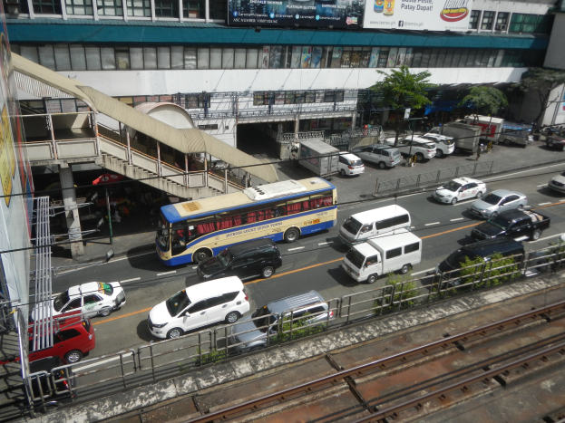 Eine belebte Stadtstraße mit Fahrzeugen, Gebäuden, Grünflächen und verschiedenen städtischen Elementen.
