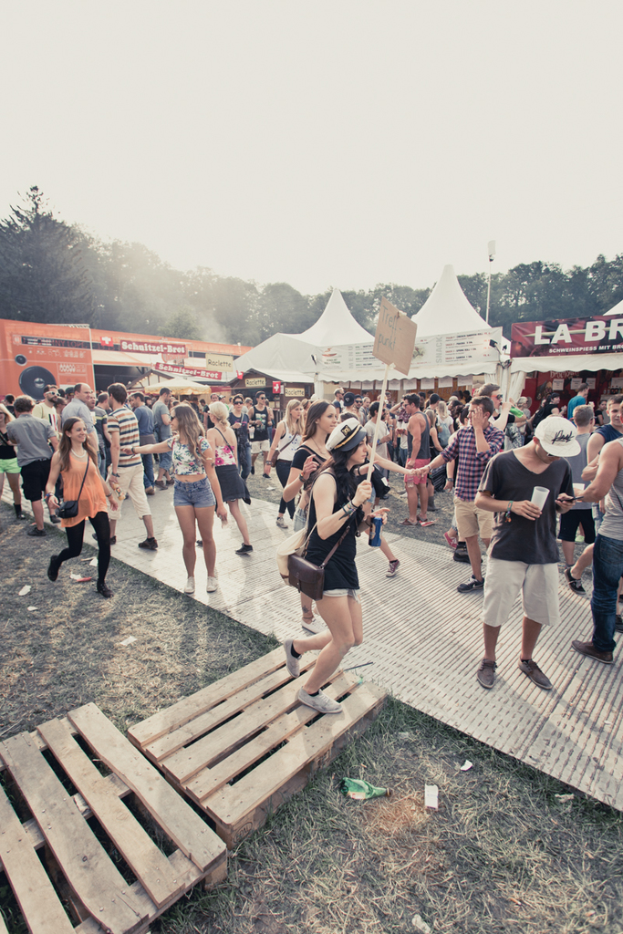 Menschen stehen und gehen auf einer Straße mit Zelten, Bäumen und einem Himmel im Hintergrund.