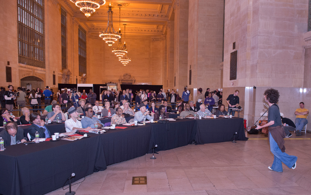 Eine Konzertszene mit einer Menschenmenge, einem Mann, der auf der rechten Seite Gitarre spielt und singt, einer Wand und Fenstern im Hintergrund, Lichtern oben und einem Boden unten.