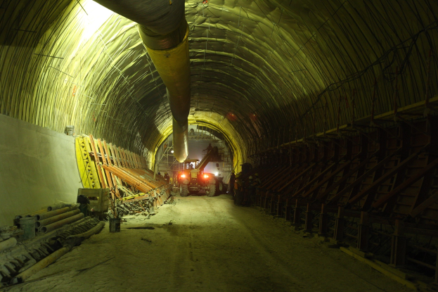 Baustelle mit einem großen Tunnel, Fahrzeugen, verstreuten hölzernen Gegenständen, Rohren und einer Wand auf der linken Seite, beleuchtet von Deckenlampen.