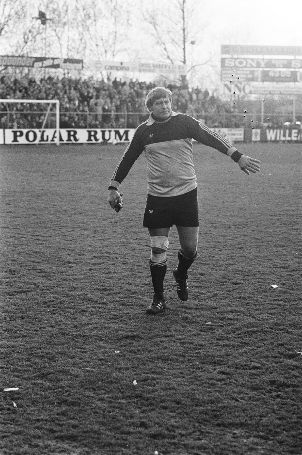 Ein Mann in einem Fußballtrikot steht auf einem Feld, umgeben von einer Menge, einem Tor, Tafeln und Bäumen unter einem klaren Himmel.