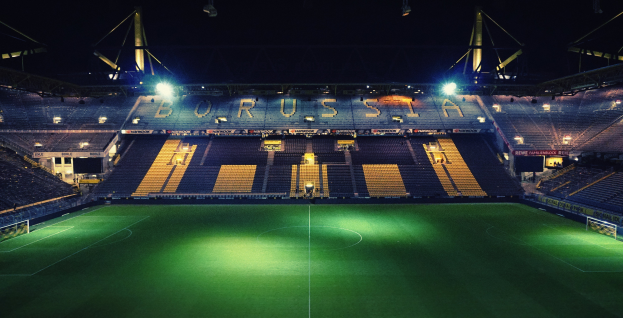 Ein Fußballfeld bei Nacht, beleuchtet von hellen Stadionlampen, mit einigen Menschen auf den Tribünen und grünem Rasen auf dem Boden.