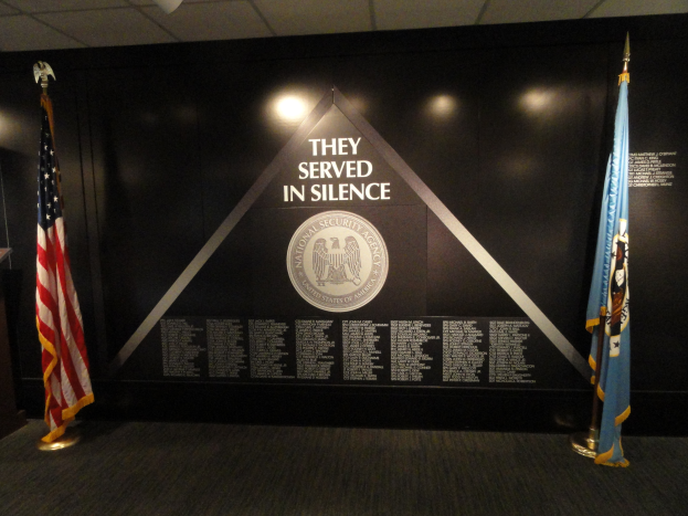 Eine Wand mit einem Schild, auf dem "Sie dienten still" steht, und zwei Fahnen davor, ein Podium mit einem Mikrofon links daneben und Lichter mit einem Logo im Hintergrund.