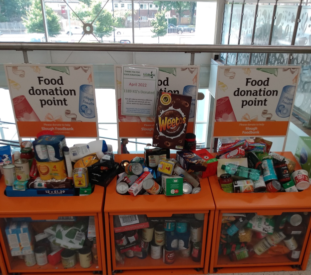 Orange Schübe mit Lebensmittelspenden gefüllt, darunter Flaschen, Dosen und Kisten, mit informativen Schildern im Hintergrund, vor einem Fenster mit Blick auf Fahrzeuge, Bäume, Gebäude und Himmel.