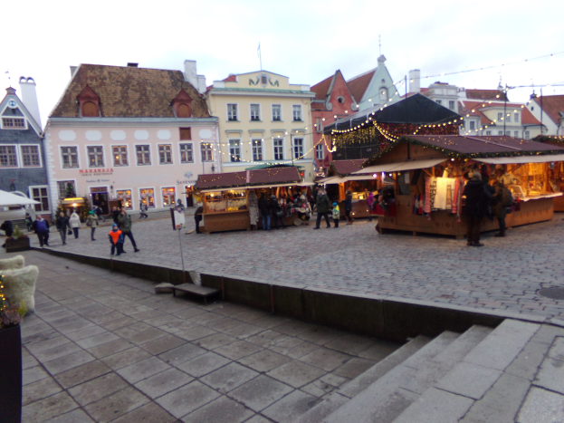 Ein belebter Weihnachtsmarkt in Tallinn, Estland mit Menschen um geschmückte Stände, festlicher Beleuchtung, bewölktem Himmel, Gebäuden im Hintergrund, Treppen und Topfpflanzen.