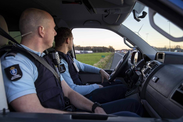 Zwei Polizeibeamte sitzen in einem Auto auf einer Straße mit Gras, Bäumen und Pfählen im Hintergrund unter einem klaren blauen Himmel.