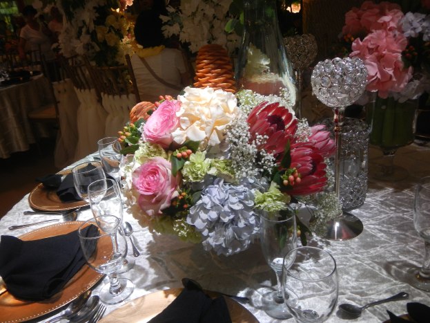 Ein formeller Esstisch mit Tellern, Besteck, Gläsern, Servietten und einem Blumenstrauß in der Mitte, umgeben von Stühlen und besetzten Tischen im Hintergrund.