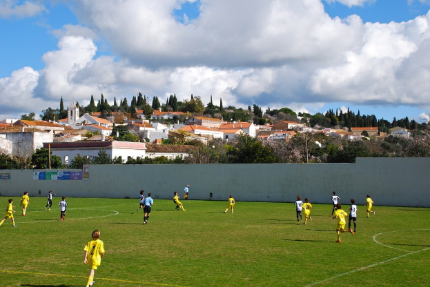 Eine Gruppe von Menschen spielt ein Spiel im Freien mit Plakaten an einer nahen Wand, Bäumen, Strommasten, Häusern und Wolken im Hintergrund.