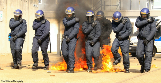 Menschen in Helmen vor einem Feuer mit verschiedenen Gegenständen auf dem Boden, Gebäuden im Hintergrund, einem Fahrzeug und einem Plakat und einer Tafel an der linken Wand, mit Text am unteren Bildrand.