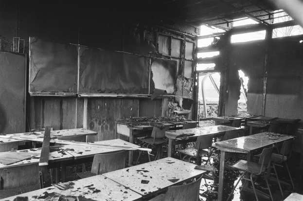 Schwarzes und weißes Foto eines Klassenzimmers mit verbrannten Wänden, zerbrochenen Fenstern und verstreuten Tischen und Stühlen.