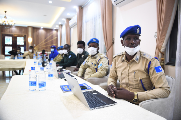 Eine Gruppe uniformierter Polizisten sitzt um einen Tisch mit Laptops, Flaschen, Büchern und anderen Gegenständen in einem Raum mit Lampen, einem Kronleuchter, Fenstern mit Vorhängen und einer Klimaanlage.