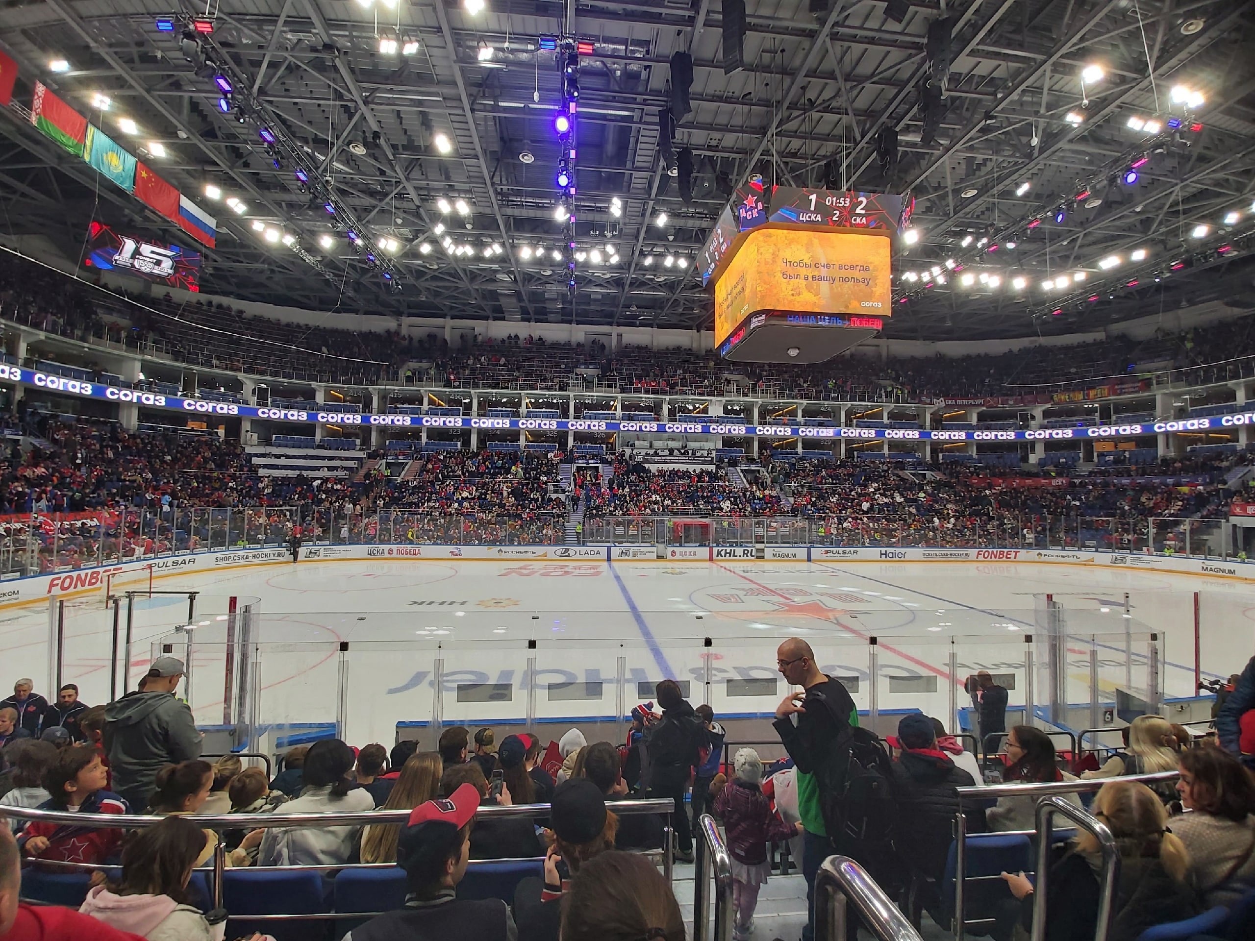 Eine große Menschenmenge schaut einem Hockey-Spiel in einer hell erleuchteten Arena mit Zuschauern, die auf und um eine von einem Zaun umgebene Eisfläche sitzen und stehen, unter von der Decke hängenden Bannern.