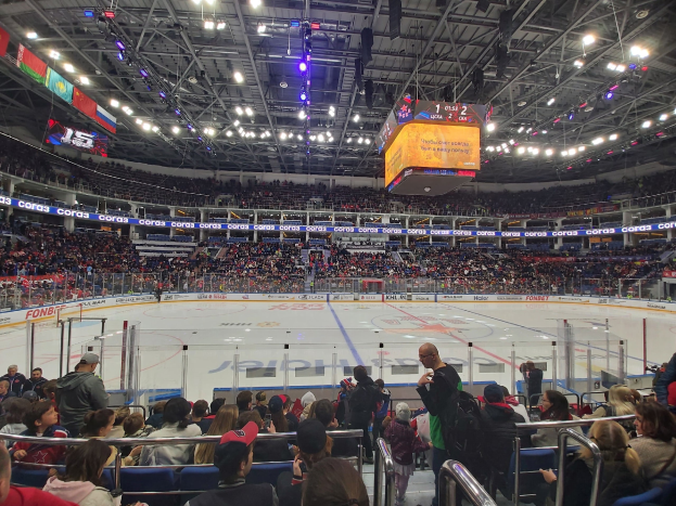 Eine große Menschenmenge schaut einem Hockey-Spiel in einer hell erleuchteten Arena mit Zuschauern, die auf und um eine von einem Zaun umgebene Eisfläche sitzen und stehen, unter von der Decke hängenden Bannern.