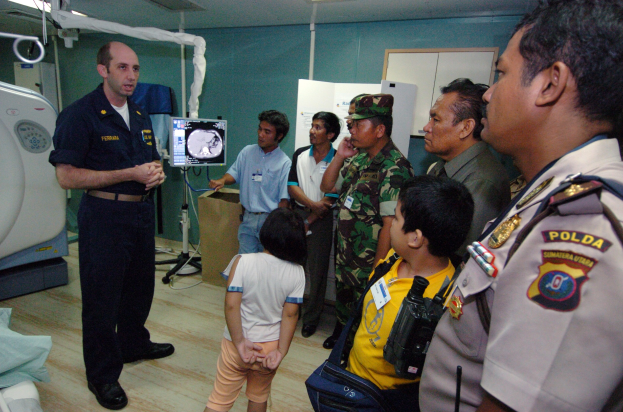 Gruppe von Menschen in einem Krankenhauszimmer mit einem Mann, der ein Mikrofon hält, einem Bett mit einer Decke darauf links, medizinischen Geräten im Hintergrund, einem Röntgenmaschinenständer, einer Texttafel, einer Tür, einer Wand und Deckenleuchten.