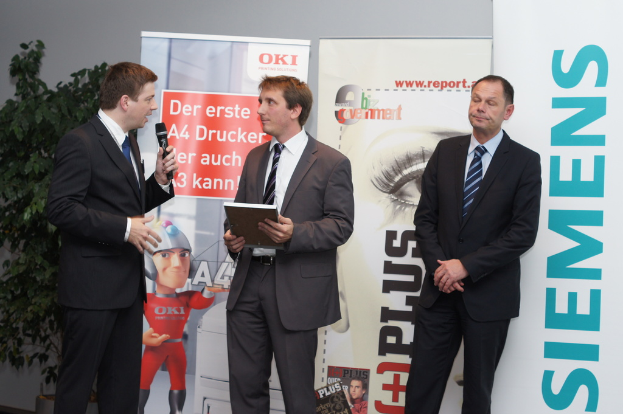 Drei Personen stehen auf einer Bühne, eine hält ein Mikrofon, eine andere ein Buch, mit Bannern und einer Pflanze dahinter.