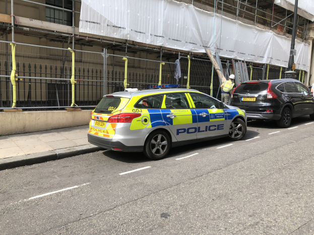 Polizeiauto auf einer Straße mit zwei Helikopterpiloten und einem weiß verhüllten Gebäude im Hintergrund, wahrscheinlich nach einer Messerstecherei in London.