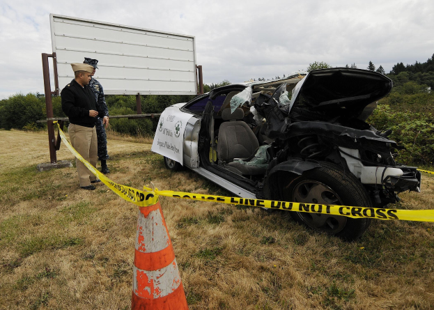 Ein Polizeibeamter in Uniform steht neben einem schwer beschädigten Auto am Straßenrand mit eingedellter Haube und zersplitterten Fenstern unter einem bewölkten Himmel.