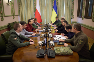 Gruppe von Männern in formeller Kleidung, die um einen Holztisch mit Mikrofonen und Dokumenten sitzen und in einem Raum mit Flaggen und Wandbeleuchtung diskutieren.