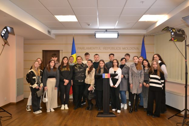 Eine Gruppe von Menschen steht vor einem Podium mit Mikrofon, umgeben von Fahnen, einer Wand mit dem Text "Moldova Youth Congress 2018", einer T├╝r, einem Dach mit Deckenleuchten und St├Ąnden mit Lichtern.