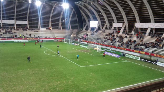 Ein Fußballspiel in einem großen Stadion mit Spielern auf dem Boden, einem Tor, eingezäunten Seitenlinien, Bannern, einem Display, einem Dach mit Metallstangen und Deckenlampen, unter einem bewölkten Himmel.