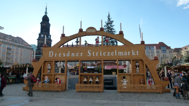 Ein lebendiger Weihnachtsmarkt in Dresden, Deutschland, mit Menschen, die herumlaufen, einige mit Taschen, und einem Bogen mit Text darauf, vor dem Hintergrund von Bäumen, Gebäuden mit Fenstern und einem bewölkten Himmel.