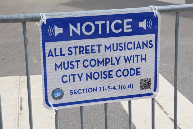 Anschlagtafel mit einem Schild, auf dem "Anschlag: Alle Straßenmusiker müssen der Stadtlärmverordnung Folge leisten" steht, an einem Zaun befestigt, mit einer Straße im Hintergrund.