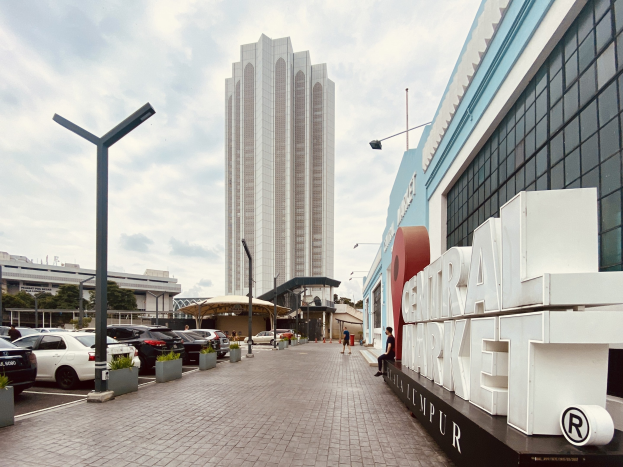 Ein belebter zentraler Markt in Kuala Lumpur, Malaysia, mit hohen Gebäuden, Fahrzeugen, Fußgängern, Straßenmöbeln und Bäumen unter einem bewölkten Himmel.