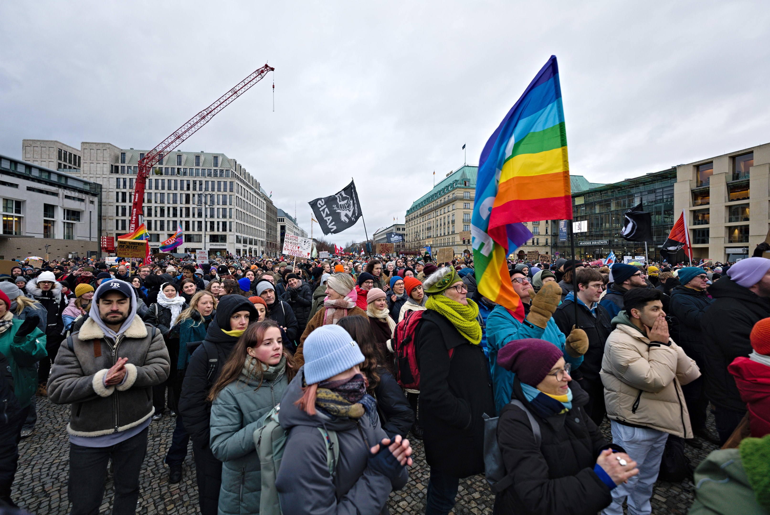 Große Gruppe von Menschen mit LGBTQ+-Rechten-Schildern während eines Marsches vor einem Gebäude mit einem Kran und einem bewölkten Himmel.